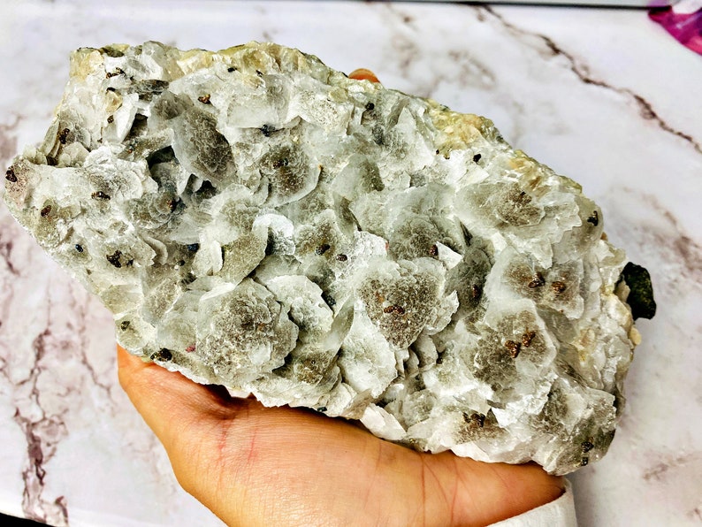 May include: A large, white, and gray crystal cluster with small, dark brown inclusions. The crystal is rough and has a natural, unpolished look.