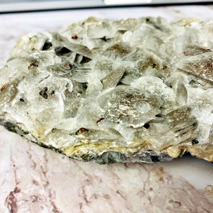 May include: A close-up of a white, layered, and sparkly mineral specimen with small, dark brown flecks throughout. The mineral is on a light brown, marbled surface.