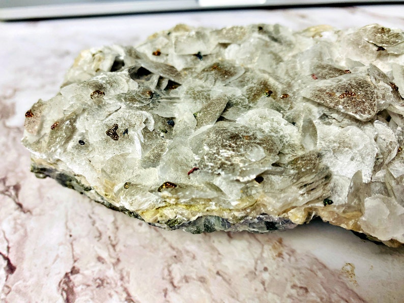 May include: A close-up of a white, layered, and sparkly mineral specimen with small, dark brown flecks throughout. The mineral is on a light brown, marbled surface.