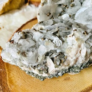 May include: A cluster of white and light pink crystals with small, shiny gold pyrite crystals embedded throughout. The crystals are arranged in a rough, natural formation and are displayed on a wooden slice.