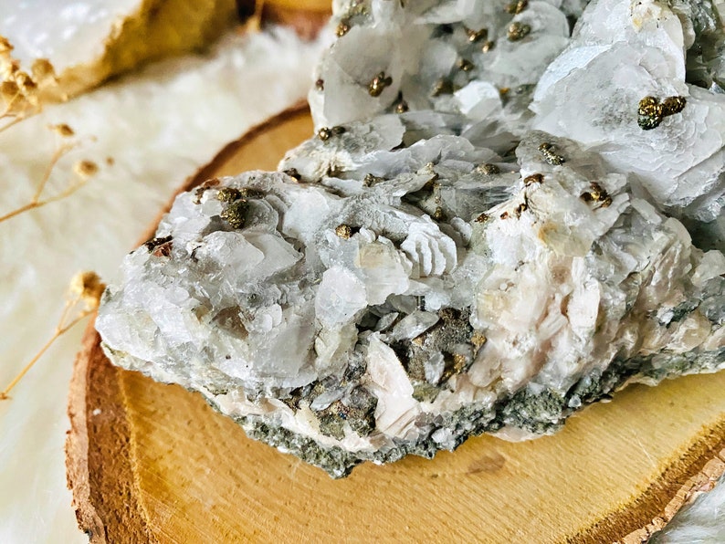 May include: A cluster of white and light pink crystals with small, shiny gold pyrite crystals embedded throughout. The crystals are arranged in a rough, natural formation and are displayed on a wooden slice.