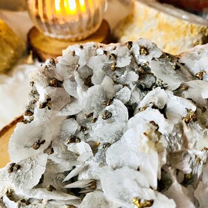 May include: Close-up of a white and gray crystal cluster with small, gold-colored pyrite crystals embedded throughout. The crystal cluster is on a wooden surface.