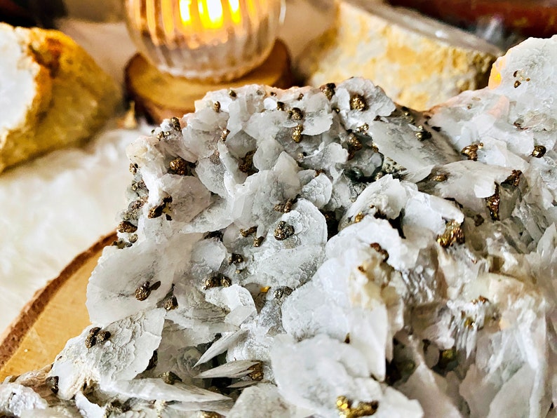 May include: Close-up of a white and gray crystal cluster with small, gold-colored pyrite crystals embedded throughout. The crystal cluster is on a wooden surface.