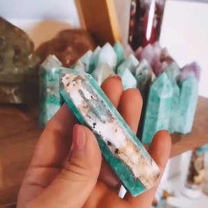 May include: A green and white crystal point, also known as a wand, is held in a hand. The crystal is a light green color with white streaks running through it. The crystal is smooth and polished.