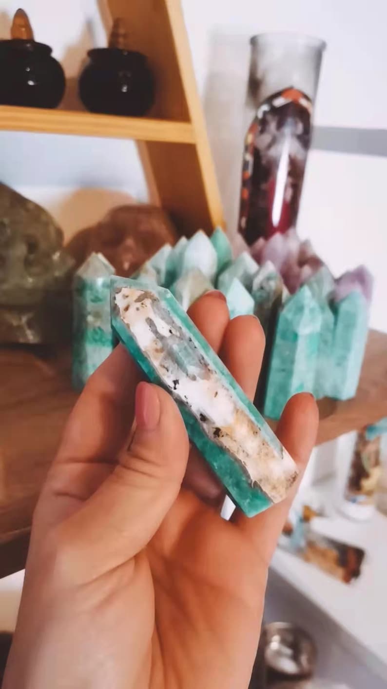 May include: A green and white crystal point, also known as a wand, is held in a hand. The crystal is a light green color with white streaks running through it. The crystal is smooth and polished.