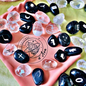 May include: A collection of black and clear tumbled stones with various symbols etched on them, including a crescent moon, a sun, a cross, a star, and a bird. The stones are in a pink pouch with the text "Les Cristaux d'Ana" printed on it.