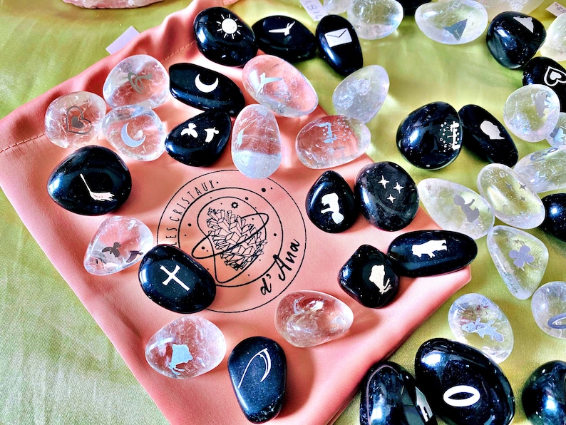 May include: A collection of black and clear tumbled stones with various symbols etched on them, including a crescent moon, a sun, a cross, a star, and a bird. The stones are in a pink pouch with the text "Les Cristaux d'Ana" printed on it.
