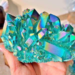 Peut inclure: Un amas de cristaux irisés aux tons de turquoise, de turquoise et de violet. Les cristaux ont un aspect chatoyant et métallique, avec des facettes angulaires et tranchantes. Les cristaux sont tenus dans une main, mettant en valeur leur taille et leurs détails complexes. Le thème général est naturel et décoratif.
