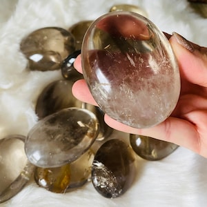 Puede incluir: Una colección de piedras de cuarzo ahumado lisas de forma ovalada. Las piedras son translúcidas y tienen un color marrón claro.