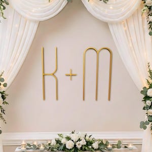 Puede incluir: Decoración de boda con iniciales doradas y un signo más en una pared crema. Cortinas blancas y arreglos florales con rosas y vegetación enmarcan la escena. Una mesa con velas y flores completa el elegante escenario.