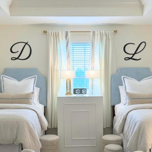 May include: Two light blue upholstered headboards with white bedding and beige lumbar pillows. Black cursive letters D and L are mounted on the wall above each bed. A white nightstand sits between the beds with a digital clock showing 10:18.