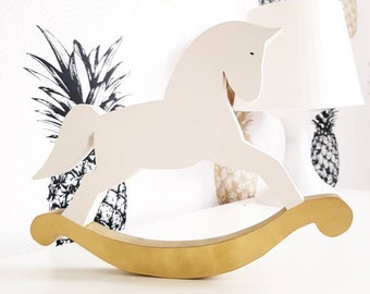 May include: A white wooden rocking horse with a gold base. The horse is a simple silhouette with a rounded head and a long, flowing mane. The base is a curved piece of wood that allows the horse to rock back and forth.