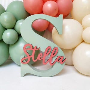 Könnte beinhalten: Ein großer, hell salbeigrüner Holzbuchstabe S mit dem Namen Stella in einer kursiven, korallenfarbenen Schriftart auf der Vorderseite. Der Buchstabe ist von einer Ansammlung von Luftballons in Salbeigrün, Koralle und Creme umgeben.