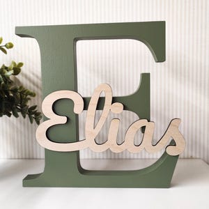 Op de afbeelding: Een grote, olijfgroene houten letter "E" met de naam "Elias" in een lichtere houttint. De naam staat in een cursief lettertype en is over de letter geplaatst. De achtergrond is een wit oppervlak met een gestreepte muur.