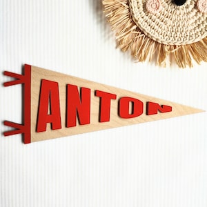 Könnte beinhalten: Holz-Wimpel mit dem Namen "ANTON" in roten Buchstaben. Der Wimpel ist hellbraun und hat eine dreieckige Form. Im Hintergrund befindet sich ein dekorativer Löwenkopf aus geflochtenem Stroh.