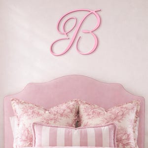 Peut inclure: Une chambre rose avec une grande lettre "B" rose fixée au mur au-dessus d'un lit. Le lit a une tête de lit rose, des oreillers à motifs floraux et un traversin à rayures. La literie a un motif floral assorti.