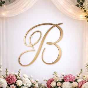 Peut inclure: Une grande lettre "B" dorée est au centre, sur fond blanc. L'écriture élégante est entourée d'arrangements floraux dans les tons de rose, blanc et crème. Un tissu blanc transparent et des guirlandes lumineuses ajoutent une touche romantique.