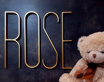 May include: Large gold-colored metal letters spelling the word ROSE mounted on a dark textured surface, positioned next to a fluffy light brown teddy bear wearing a brown satin ribbon tied in a bow around its neck.