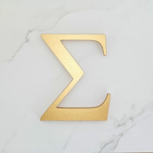 Könnte beinhalten: Ein goldfarbener griechischer Buchstabe Sigma aus Holz, der auf einer weißen Marmoroberfläche liegt. Der Buchstabe hat ein schlichtes, geometrisches Design mit einer flachen, matten Oberfläche. Sigma wird oft in Mathematik und Statistik verwendet.