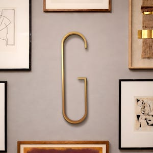 May include: A large, gold-colored metal letter G mounted on a light gray wall. The letter features a tall, elongated, and minimalist sans-serif design with a smooth, matte finish, positioned centrally among several framed art pieces.