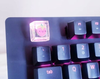 May include: A black keyboard with pink backlighting. The keyboard has a keycap with a strawberry design. The keycap is clear and the strawberry is pink with a yellow stem and green leaves. The strawberry has a happy face.