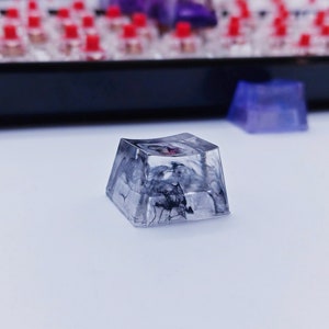 May include: A clear, square-shaped keycap with a black and white marbled design. The keycap has a slight curve to it and is made of resin.