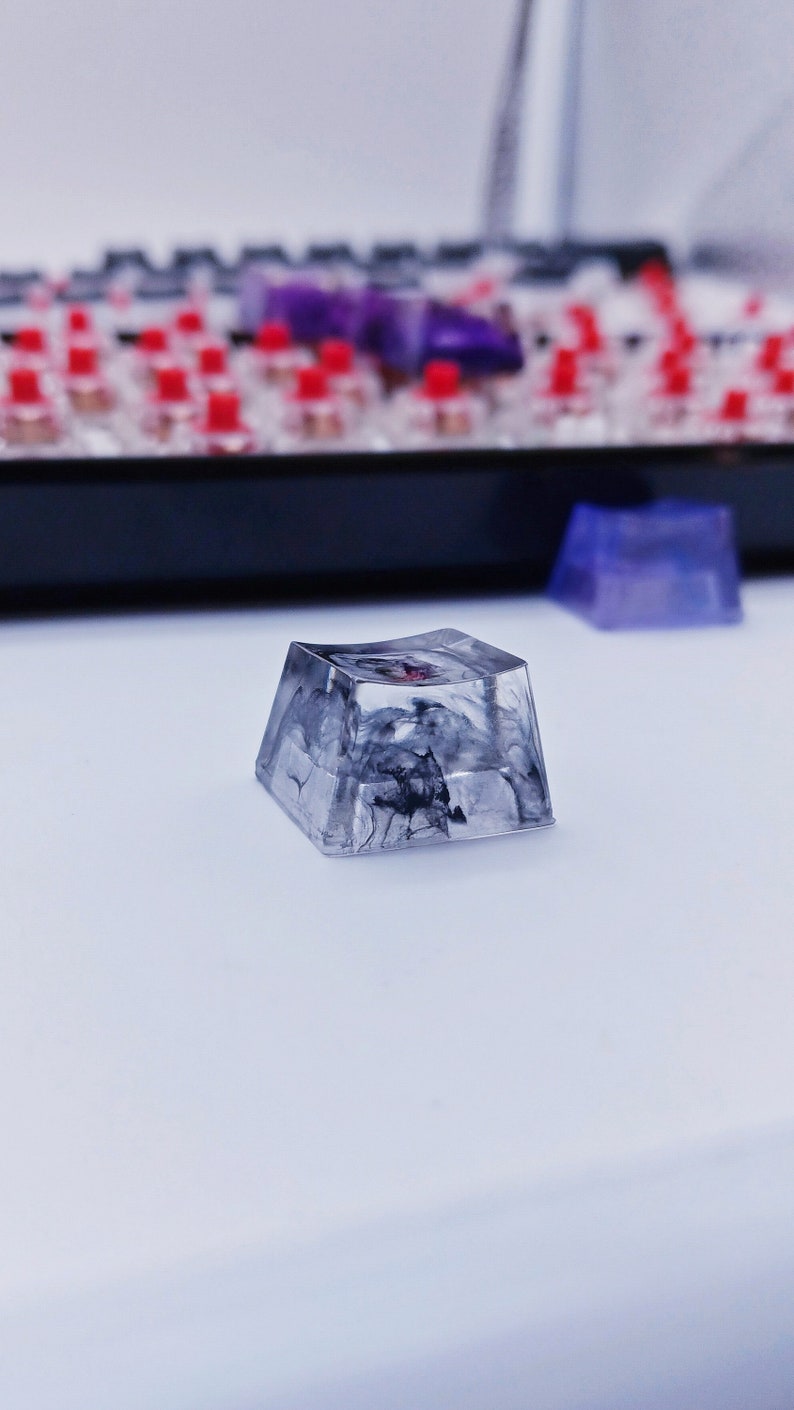May include: A clear, square-shaped keycap with a black and white marbled design. The keycap has a slight curve to it and is made of resin.