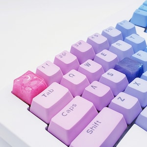 Könnte beinhalten: Eine rosa Tastatur mit einer benutzerdefinierten Tastenkappe mit Mond- und Sternenmotiv. Die Tastatur hat einen Farbverlauf, der von Hellrosa zu Lila übergeht.