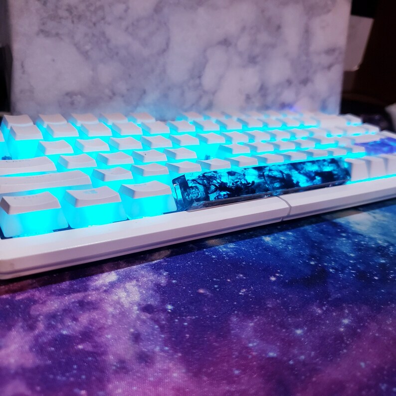 May include: A white mechanical keyboard with blue backlighting and a custom blue and black resin keycap. The keyboard is sitting on a purple and blue galaxy-themed mousepad.