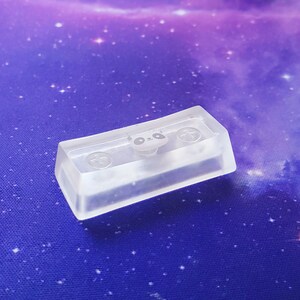 May include: A clear plastic keycap with a panda face design and two plus signs. The keycap is on a purple and white background.