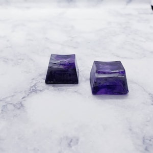 May include: Two purple resin keycaps with a marbled effect. The keycaps are square shaped and have a slightly curved top.