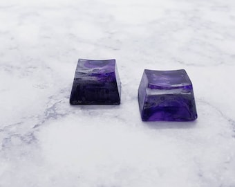 May include: Two purple resin keycaps with a marbled effect. The keycaps are square shaped and have a slightly curved top.