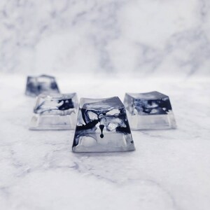 May include: Four clear resin keycaps with black marbling. The keycaps are square shaped and have a slightly rounded top.