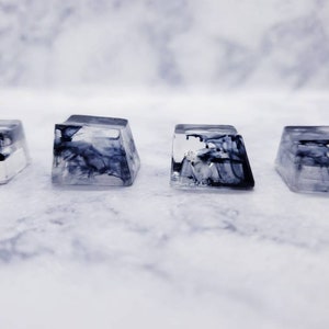 May include: Four clear resin keycaps with black and white marbled designs. The keycaps are square and have a slightly rounded top.