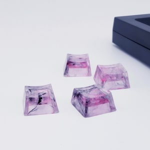 May include: Four translucent pink and black keycaps with a marbled design. The keycaps are square shaped and have a slight curve on the top.