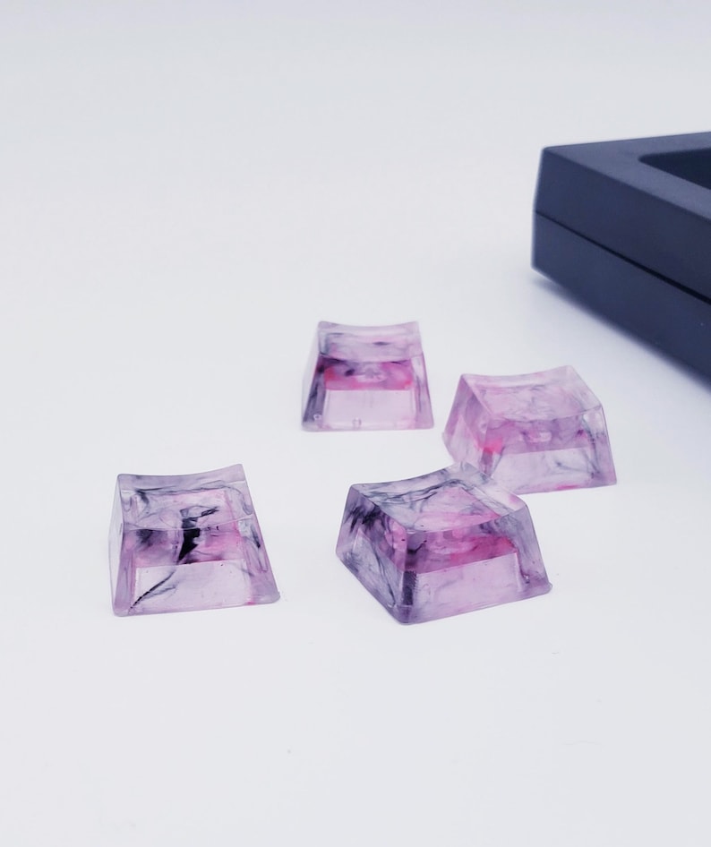 May include: Four translucent pink and black keycaps with a marbled design. The keycaps are square shaped and have a slight curve on the top.