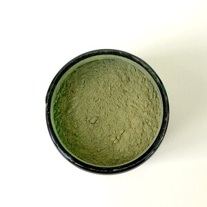 Pu&ograve; includere: Una vista ravvicinata di un piccolo contenitore rotondo nero pieno di una polvere verde chiaro. La polvere &egrave; liscia e distribuita uniformemente in tutto il contenitore.
