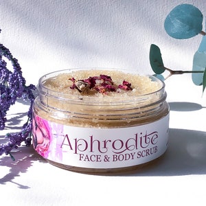 May include: A clear glass jar filled with a light brown sugar scrub with dried rose petals on top. The jar has a white label with the text "Aphrodite FACE & BODY SCRUB".