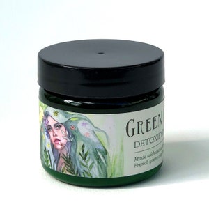 Pu&ograve; includere: Un barattolo verde con un coperchio nero e un'etichetta che dice "GREEN DETOXIFY" e "Realizzato con argilla verde francese naturale". L'etichetta presenta anche un'illustrazione ad acquerello di una donna con i capelli verdi e fiori.