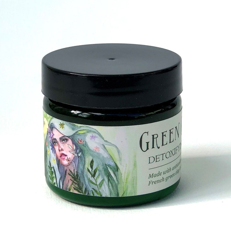 Pu&ograve; includere: Un barattolo verde con un coperchio nero e un'etichetta che dice "GREEN DETOXIFY" e "Realizzato con argilla verde francese naturale". L'etichetta presenta anche un'illustrazione ad acquerello di una donna con i capelli verdi e fiori.
