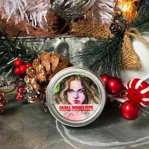 May include: A silver tin with a red and white label that reads "Cocoa Peppermint Lip Balm". The label features a watercolor illustration of a woman with long brown hair wearing a wreath of green leaves. The tin is surrounded by red and white candy canes and red berries.