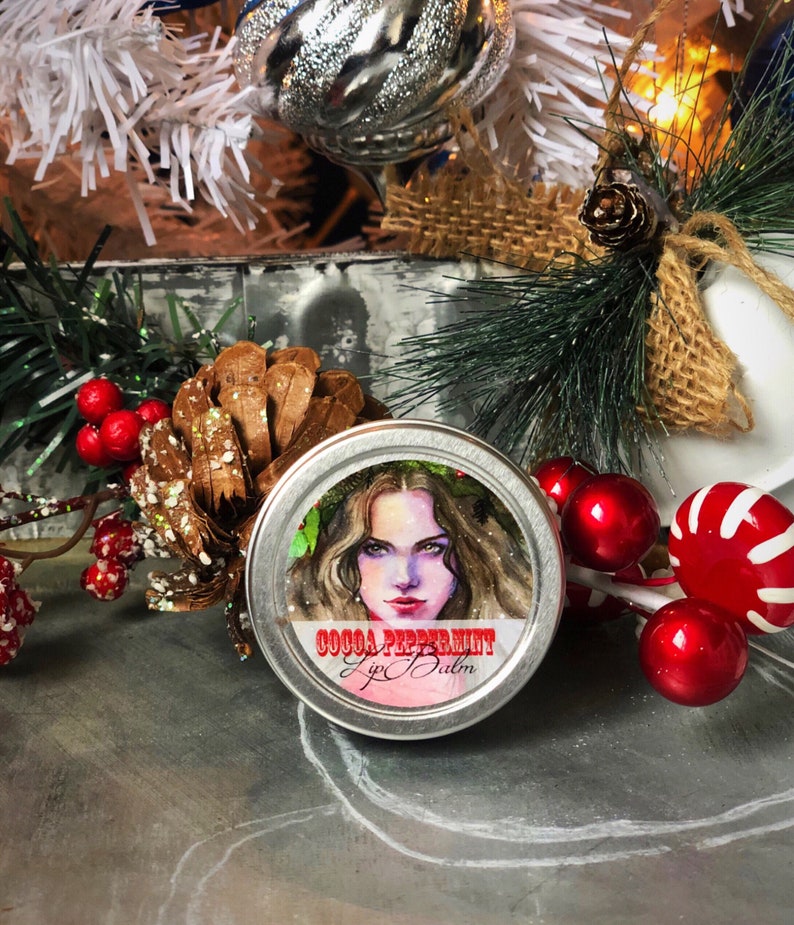 May include: A silver tin with a red and white label that reads "Cocoa Peppermint Lip Balm". The label features a watercolor illustration of a woman with long brown hair wearing a wreath of green leaves. The tin is surrounded by red and white candy canes and red berries.