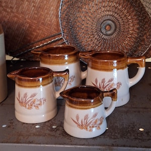 May include: A set of four ceramic creamers in varying sizes, each featuring a cream-coloured body with a brown glazed rim and a brown wheat stalk pattern printed on the front. Each creamer has a small pouring spout and a single side handle.