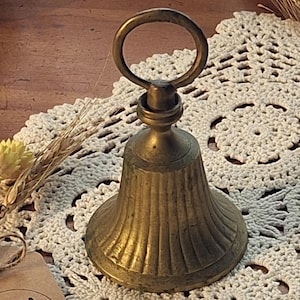 May include: A brass bell with a fluted body and a circular handle at the top. A small brown paper tag is attached to the bell, featuring the printed text "Joy On the Farm" along with a small wheat graphic and additional small text.
