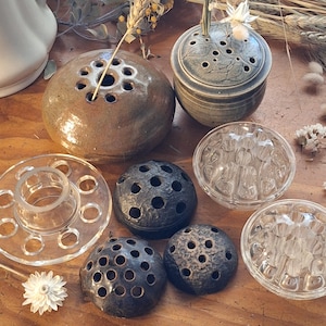 May include: A collection of seven flower frogs in various materials and shapes. The set includes two ceramic vessels with perforated lids, three dark textured metal domes with multiple holes, and two clear glass floral arrangers with circular openings.