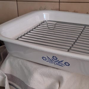 May include: A rectangular white ceramic roasting pan featuring a blue floral pattern on the side. The pan includes a removable silver-toned metal wire rack that fits inside the basin. The exterior surface is smooth and glossy with a clean finish.