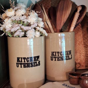 May include: Two beige ceramic kitchen utensil holders with the words "KITCHEN UTENSILS" in black. One holds white flowers, the other wooden spoons. Additional small brown ceramic pots and a knife are visible.