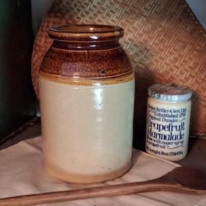 May include: A traditional stoneware jar with a brown top and a cream-coloured body. A small jar of grapefruit marmalade and a wooden spoon are on a cloth. A woven basket is in the background.