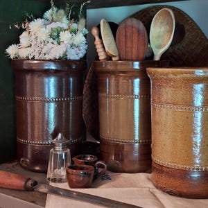 May include: A kitchen still life with three brown ceramic utensil pots, one holding dried flowers. Wooden spoons and other kitchen tools are visible. A small glass salt shaker and miniature ceramic cups are also present.