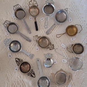 May include: A selection of vintage tea strainers in various shapes and sizes, made of metal. Some have handles, others are round or teapot-shaped. The strainers are displayed on a white, textured surface.